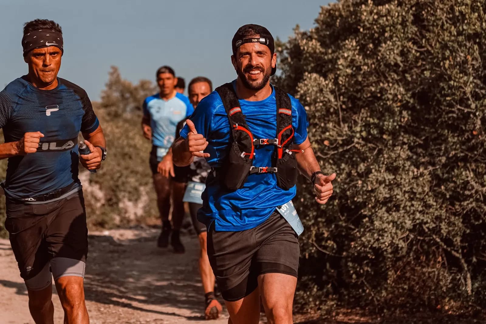 Un coureur souriant lève le pouce sur un sentier de trail, suivi par d’autres participants